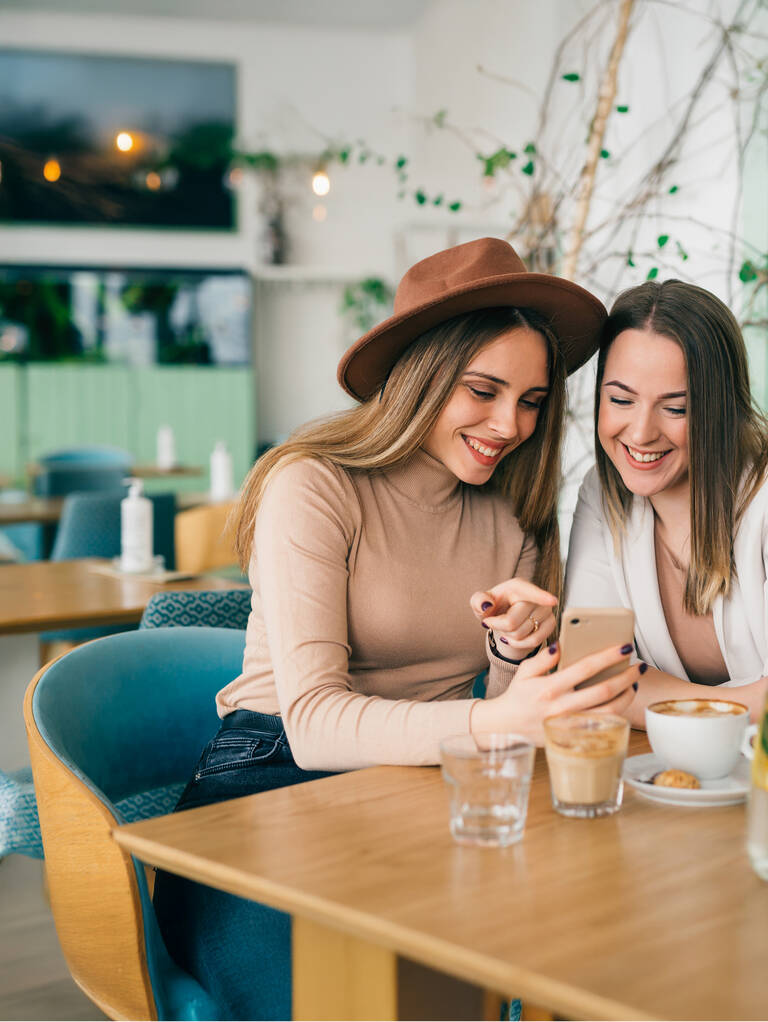 Zwei Frauen sitzen lächelnd nebeneinander in einem Café und schauen auf ihr Handy
