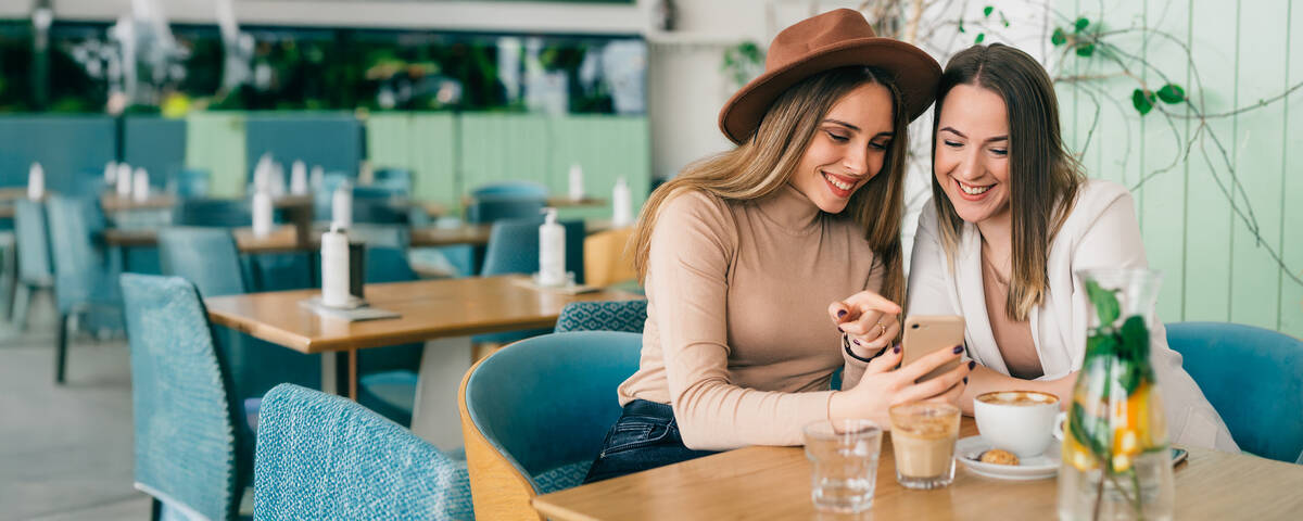 Zwei Frauen sitzen lächelnd nebeneinander in einem Café und schauen auf ihr Handy