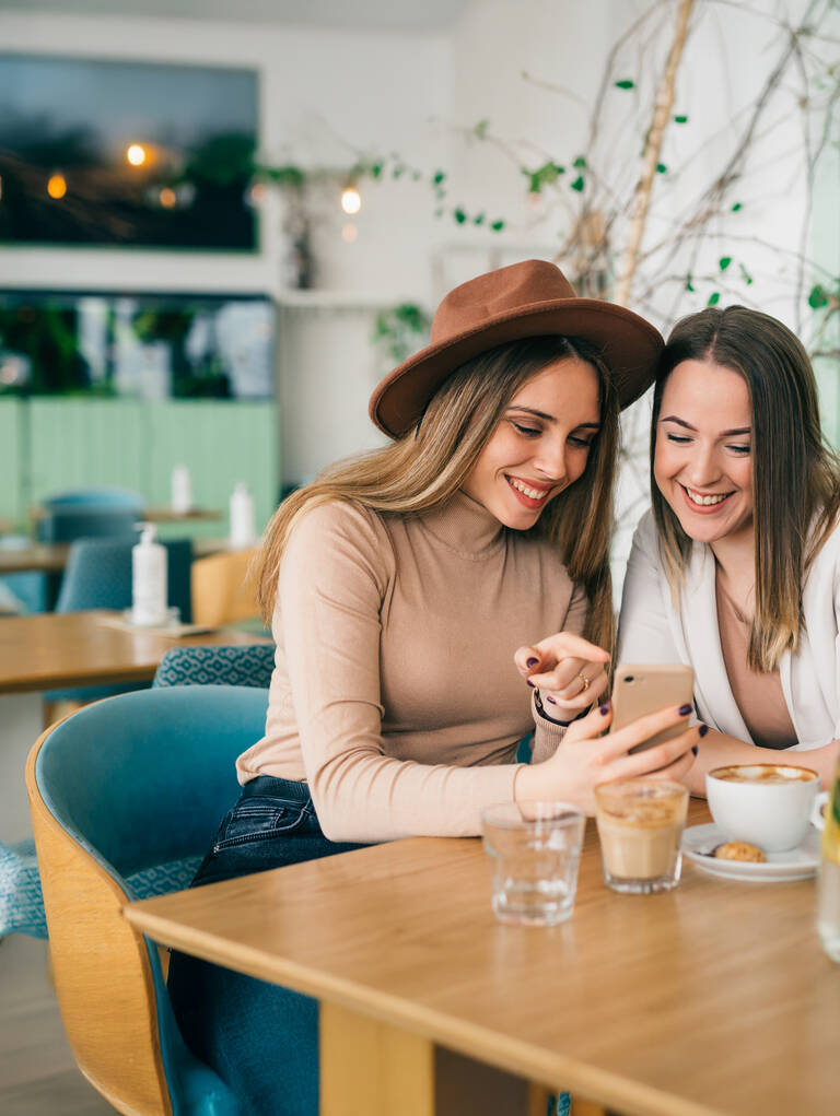 Zwei Frauen sitzen lächelnd nebeneinander in einem Café und schauen auf ihr Handy