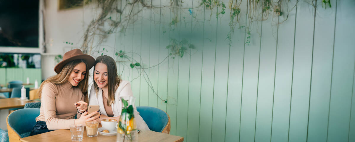 Zwei Frauen sitzen lächelnd nebeneinander in einem Café und schauen auf ihr Handy